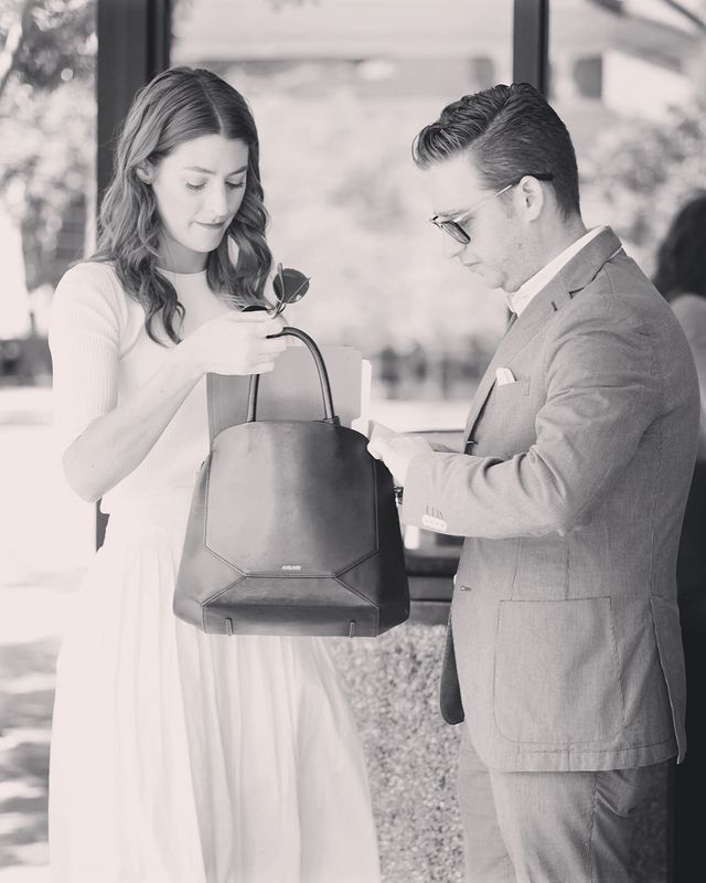 Trevor Einhorn in a formal suit putting stuff in his wife's purse.